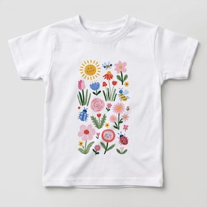 Puede incluir: Una camiseta blanca de manga corta con un estampado gráfico central que muestra un sol amarillo sonriente, varias flores coloridas en tonos rosa, rojo y azul, dos abejas y dos mariquitas dispuestas en un patrón vertical en la parte frontal.