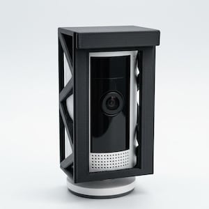 May include: A black security camera cover with a geometric design. The cover surrounds a white and black security camera with a lens and speaker holes. The camera sits on a white base, all against a white background.