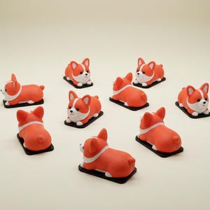 May include: A collection of orange and white corgi dog figurines. Each figurine is lying down on a black rectangular base. The corgis have white faces, chests, and paws, with orange bodies and ears.