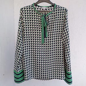 Pode incluir: Uma blusa de manga comprida com um padrão geométrico em preto, branco e verde. A blusa tem um laço no decote e acabamentos verdes na gola e punhos. A blusa está pendurada num cabide preto.