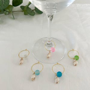 May include: Five gold-colored wine glass charms with different colored glass beads and a small seashell charm. The beads are pink, green, light blue, and dark blue.