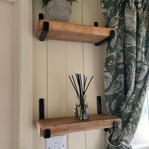 May include: Two wooden shelves with black metal brackets mounted on a white wall. The top shelf has a potted plant on it. The bottom shelf has a glass bottle with black sticks in it. A green patterned curtain is to the right of the shelves.