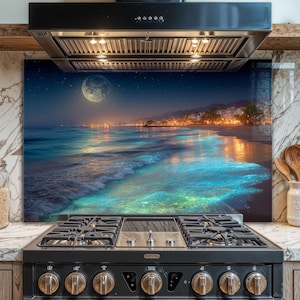 Puede incluir: Un protector de salpicaduras de cocina de vidrio con una escena de playa nocturna con luna llena, cielo estrellado y agua turquesa brillante. Montado sobre una estufa de gas negra y debajo de una campana extractora de acero inoxidable.