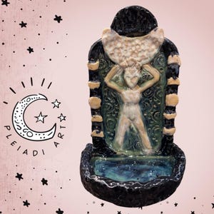 Puede incluir: Un lavabo de pared de cerámica con una figura central esculpida que sostiene una gran luna creciente sobre su cabeza. La pieza está acabada con esmaltes negro oscuro, verde profundo y crema, con fases lunares en los bordes verticales.