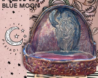 Può includere: Una ciotola altare in ceramica con finitura lucida nei toni del viola, blu e rosa. L'interno presenta un rilievo di una figura, mentre l'esterno è decorato con un motivo floreale ripetuto. Il testo ALTAR BLUE MOON è visibile.