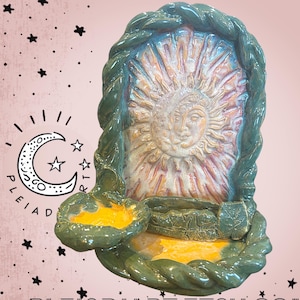 Può includere: Un porta incenso in ceramica con un rilievo centrale raffigurante un volto di sole e luna circondato da un bordo a corda intrecciata. L'oggetto include una piccola ciotola dipinta di giallo e una base piatta con vassoio testurizzato a foglia, smalto blu ottanio.