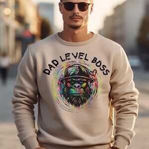May include: A beige sweatshirt with a colorful graphic of a gorilla wearing a hat and sunglasses. The text "DAD LEVEL BOSS" is printed in black around the graphic.