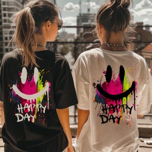 May include: Black and white t-shirts with a smiley face design. The design features a pink and yellow smiley face with paint splatter and the words "Happy Day". The shirts are short-sleeved.
