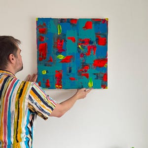 Puede incluir: Pintura de arte abstracto con una vibrante mezcla de colores. La obra presenta un lienzo cuadrado con un fondo turquesa, acentuado por pinceladas rojas y amarillas. La pintura se cuelga en una pared blanca.