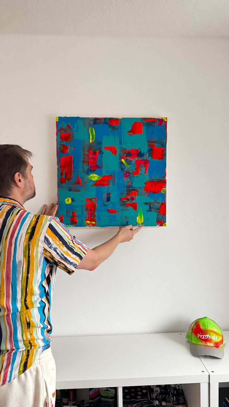 Puede incluir: Pintura de arte abstracto con una vibrante mezcla de colores. La obra presenta un lienzo cuadrado con un fondo turquesa, acentuado por pinceladas rojas y amarillas. La pintura se cuelga en una pared blanca.