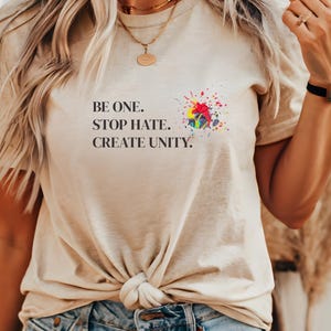 Könnte beinhalten: Ein beiges T-Shirt mit einem bunten Farbklecks-Design und dem Text "Be One. Stop Hate. Create Unity."