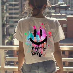 Könnte beinhalten: Ein weißes Kurzarm-T-Shirt mit einem großen, bunten Farbspritzer-Design auf der Rückseite. Die Grafik zeigt ein schwarzes, tropfendes Smiley-Gesicht und den Text "HAPPY DAY" in Schwarz, ergänzt durch ein kleines, verzerrtes Smiley-Symbol.