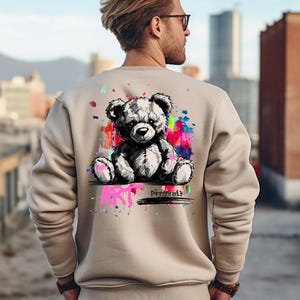 May include: Beige sweatshirt featuring a black and white teddy bear graphic with colorful paint splatter accents. The word "Prompt" is printed below the bear. The sweatshirt is a neutral color and has long sleeves.
