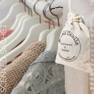 May include: A white muslin drawstring bag with a black circle logo that says "Morgan Candle Hand Poured & Crafted morgancandleshop.com". The bag is hanging on a white wooden hanger in a closet with clothes on hangers behind it.
