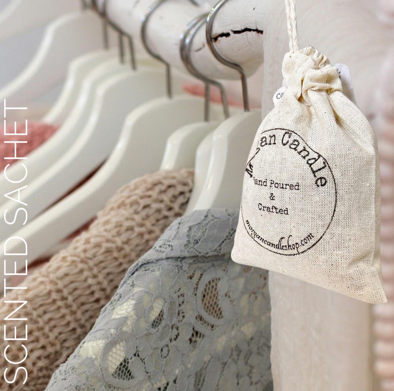 May include: A white muslin drawstring bag with a black circle logo that says "Morgan Candle Hand Poured & Crafted morgancandleshop.com". The bag is hanging on a white wooden hanger in a closet with clothes on hangers behind it.