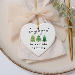 May include: A white heart-shaped ornament with the word "Engaged" in script. Below are three green pine trees, the names "Steven + Adel", and the date "25.07.2025". The ornament hangs from a gold string and is set on a plate with a napkin and a bow.
