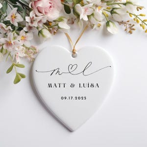 May include: White ceramic heart ornament with black calligraphy that reads "m&l" Matt & Luisa 09.17.2025.