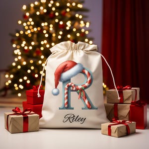 May include: A cream-colored drawstring gift bag with a Santa hat and candy cane design forming the letter "R". The name "Riley" is printed below. Several wrapped gifts with red ribbons are in the foreground, with a Christmas tree in the background.