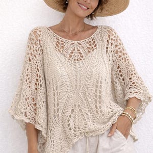 Könnte beinhalten: Eine Frau trägt ein cremefarbenes Strickoberteil mit offenem Gewebe, weiten Ärmeln und passenden Shorts. Sie trägt auch einen breitkrempigen Strohhut und goldene Armbänder. Das Outfit ist leicht und luftig, perfekt für warmes Wetter.
