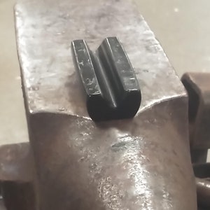 May include: A close-up of a metal anvil with a black metal anvil tool on top. The anvil is sitting on a wooden base.