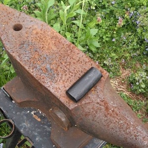 May include: A rusty metal anvil with a black rubber pad on top, sitting on a black metal stand. The anvil is outdoors on a bed of green grass.