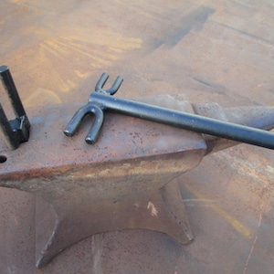 May include: A black metal tool with two U-shaped ends and a long handle, resting on a rusty anvil.