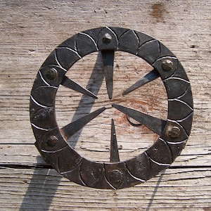 May include: A circular metal design with a decorative border of overlapping petals and a central design of six pointed spokes. The metal is dark gray and has a rustic finish.