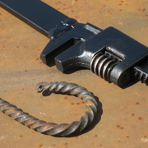May include: A black metal wrench with a serrated jaw and a twisted metal hook. The wrench is lying on a rusty metal surface.