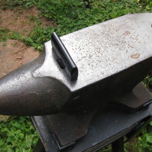 May include: A black metal anvil with a worn surface and a black metal handle on the top. The anvil is sitting on a black metal stand.