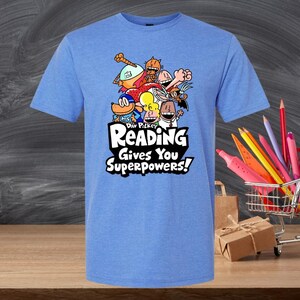 May include: A light blue t-shirt featuring a colorful cartoon graphic with the text "READING Gives You Superpowers!". The graphic includes various cartoon characters. The shirt is displayed on a wooden surface.