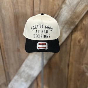Puede incluir: Gorra de béisbol crema y negra con el texto "PRETTY GOOD AT BAD DECISIONS" en negro. La gorra tiene una visera negra y un botón negro en la parte superior. La gorra se muestra en un soporte metálico.