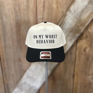 Può includere: Cappellino da baseball color crema e nero con la scritta "ON MY WORST BEHAVIOR". Il cappellino ha una visiera curva e un bottone nero sulla parte superiore. L'etichetta del marchio "OTTO" è visibile. Sfondo in legno.