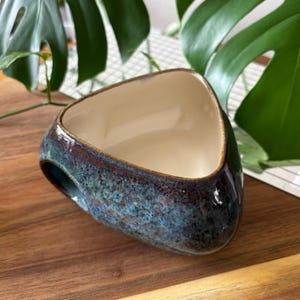 May include: A handmade ceramic bowl with a distinctive triangular form. The exterior is glazed in a mix of blue and brown, producing a textured, iridescent finish. The interior is a creamy off-white, and the bowl has a small handle.