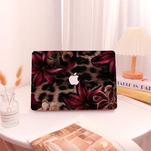 May include: A laptop case featuring a floral and leopard print design. The case has a dark background with burgundy flowers and a leopard print pattern. The Apple logo is visible in the center. The case is on a white table.