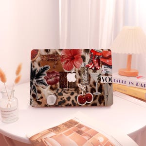 May include: A laptop case with a collage design featuring a leopard print background, red hibiscus flower, Coca-Cola can, and a red lip print. Other elements include a cocktail glass, cherries, and an apple logo.