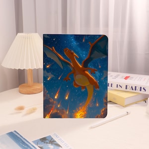 May include: A notebook featuring a vibrant illustration of a Charizard, a fiery orange dragon, soaring through a starry night sky with falling meteors. The cover has a glossy finish and a colorful design.