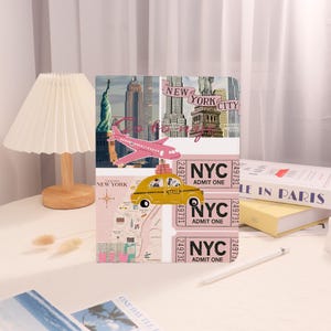 May include: A pink and white travel-themed notebook featuring illustrations of New York City landmarks, a pink airplane, and a yellow taxi. The notebook includes the text "NEW YORK CITY" and "NYC ADMIT ONE" on pink ticket-style designs.
