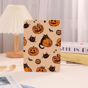 May include: A rectangular Halloween-themed decorative item featuring a repeating pattern of orange jack-o'-lanterns, black cats, paw prints, and bats on a tan background. The item is likely a phone case or a similar accessory.