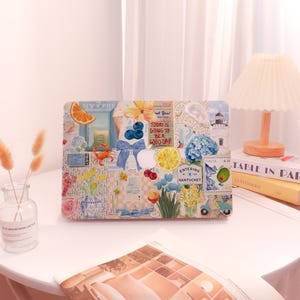 May include: A rectangular laptop case with a collage design featuring images of fruit, flowers, and nautical elements. The case has text that reads "NEW YORKER" and "TODAY IS GOING TO BE A GOOD DAY!" The case is on a white table.