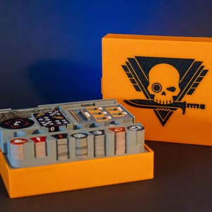 May include: An orange plastic box with a skull and crossbones logo on the lid. The box is open and contains a variety of game pieces, including dice, tokens, and cards.