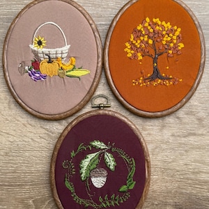 May include: Three oval embroidered wall hangings in wooden frames. One shows a basket with a sunflower, pumpkin, and cornucopia. Another depicts a tree with autumn leaves. The third shows an acorn and leaves.