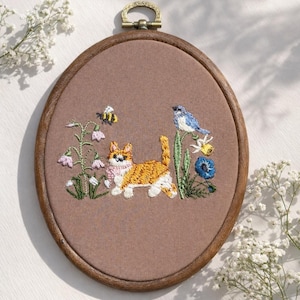 Peut inclure: Broderie ovale dans un cadre en bois. Le motif représente un chat roux et blanc avec une écharpe rose, un oiseau bleu, une abeille et des fleurs. Le fond est un tissu marron clair, avec une boucle de suspension en laiton.