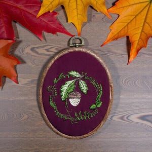 May include: Oval embroidery hoop featuring an acorn and leaf design in green and white on a deep purple background. The wooden frame is surrounded by a natural fiber. Autumn leaves in red, orange, and yellow surround the hoop.