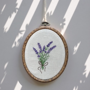 Peut inclure: Broderie ovale représentant un bouquet de lavande. La lavande violette est brodée sur un tissu blanc, nouée avec un ruban marron. Le cercle en bois est suspendu par une ficelle crème.