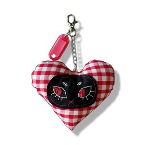 May include: A heart-shaped keyring with a red and white gingham pattern. It features a black cat face applique with red eyes and white outlines. The keyring includes a pink tag and a silver clasp, with the text "Lost Kittens" above.