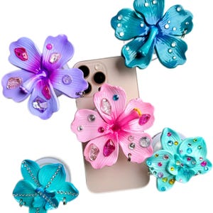 May include: Five flower-shaped phone grips in shades of pink, purple, and teal, adorned with rhinestones. The grips are attached to a phone, showcasing their decorative design and functionality. The flower-shaped grips add a touch of elegance.