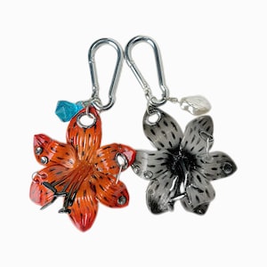 Puede incluir: Dos llaveros con forma de flor. Uno es naranja con detalles negros, el otro es negro y gris. Cada llavero tiene un mosquetón plateado y un pequeño dije: una gema azul y una perla blanca. Sobre un fondo blanco.