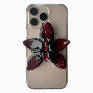 Op de afbeelding: Een donkerrode en zwarte bloemvormige telefoonhouder bevestigd aan een doorzichtige telefoonhoes. De bloem heeft vijf bloemblaadjes met een gradiënteffect, van donkerrood naar zwart. De telefoon heeft een lichtbruine kleur.