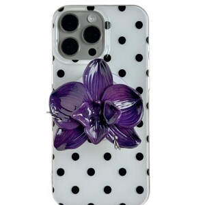 Pode incluir: Uma capa de telefone com fundo branco e bolinhas pretas. Uma orquídea roxa 3D está presa à capa. A capa do telefone tem um recorte para a câmera e um pop socket branco.