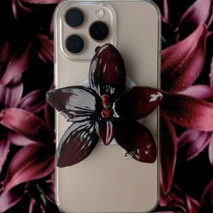 Puede incluir: Una funda de teléfono con un agarre para teléfono en forma de flor de color burdeos oscuro. La flor tiene cinco pétalos y un pequeño centro rojo. La funda del teléfono es transparente y el teléfono es de color beige claro. El fondo es un patrón floral oscuro.
